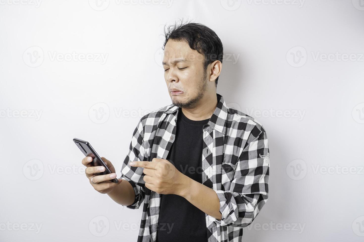 A dissatisfied young Asian man looks disgruntled wearing tartan shirt irritated face expressions holding his phone photo