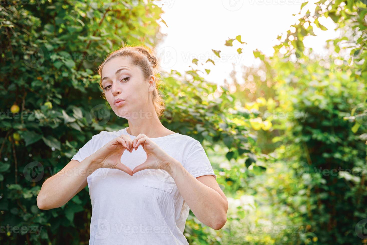 Young woman smiling outdoor. Beautiful brunete girl showing heart sign with hands on park or garden green background. Free happy woman at summertime. Freedom happiness carefree happy people concept. photo