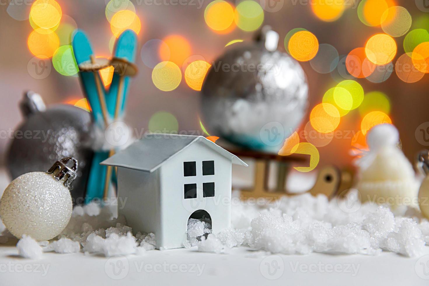 Abstract Advent Christmas Background. Toy model house and winter decorations ornaments toys and balls on background with snow and defocused garland lights. Christmas with family at home concept. photo