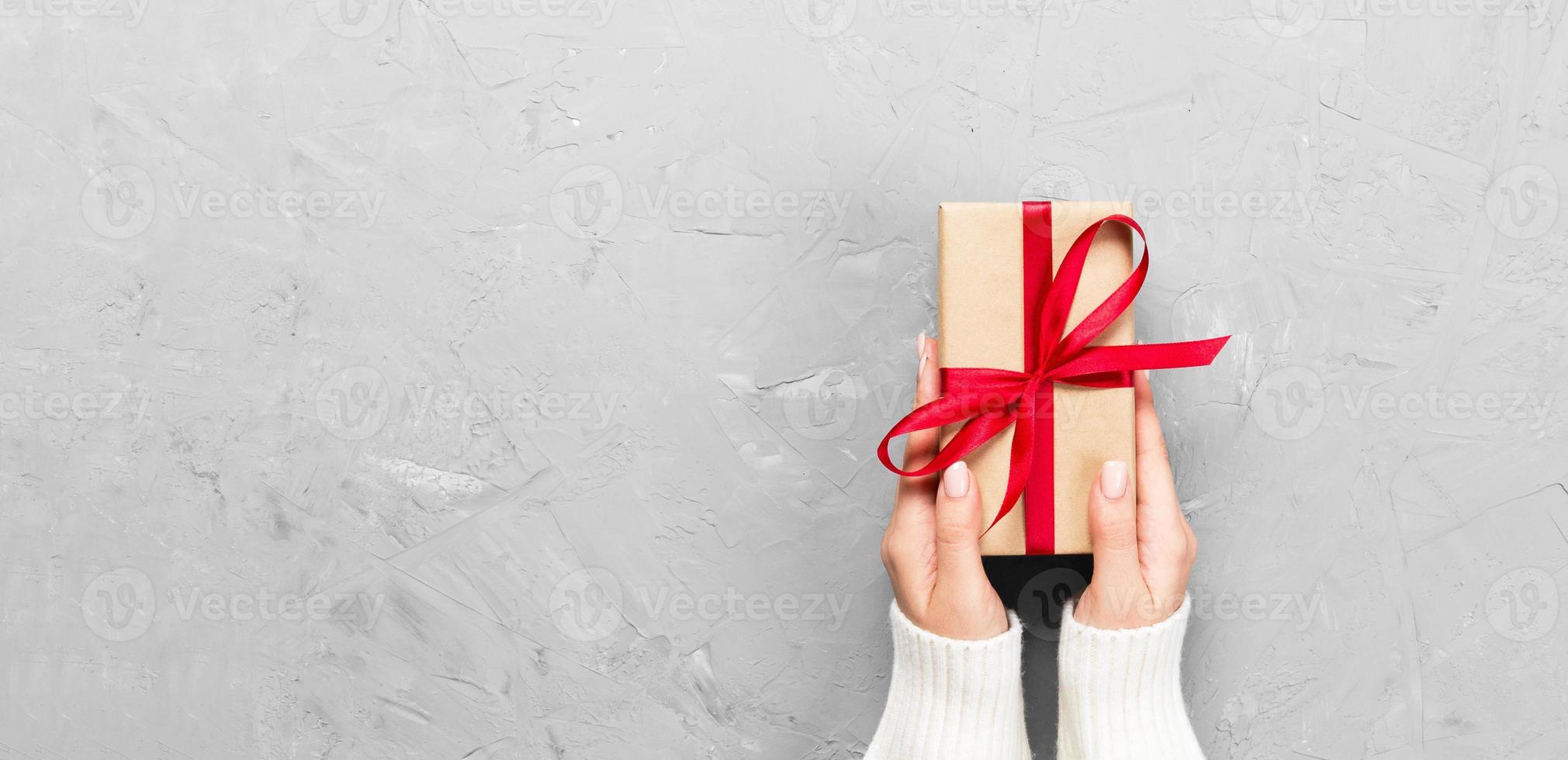 Woman hands give wrapped valentine or other holiday handmade present in paper with red ribbon. Present box, decoration of gift on white cement table, Top view banner with copy space for you design photo