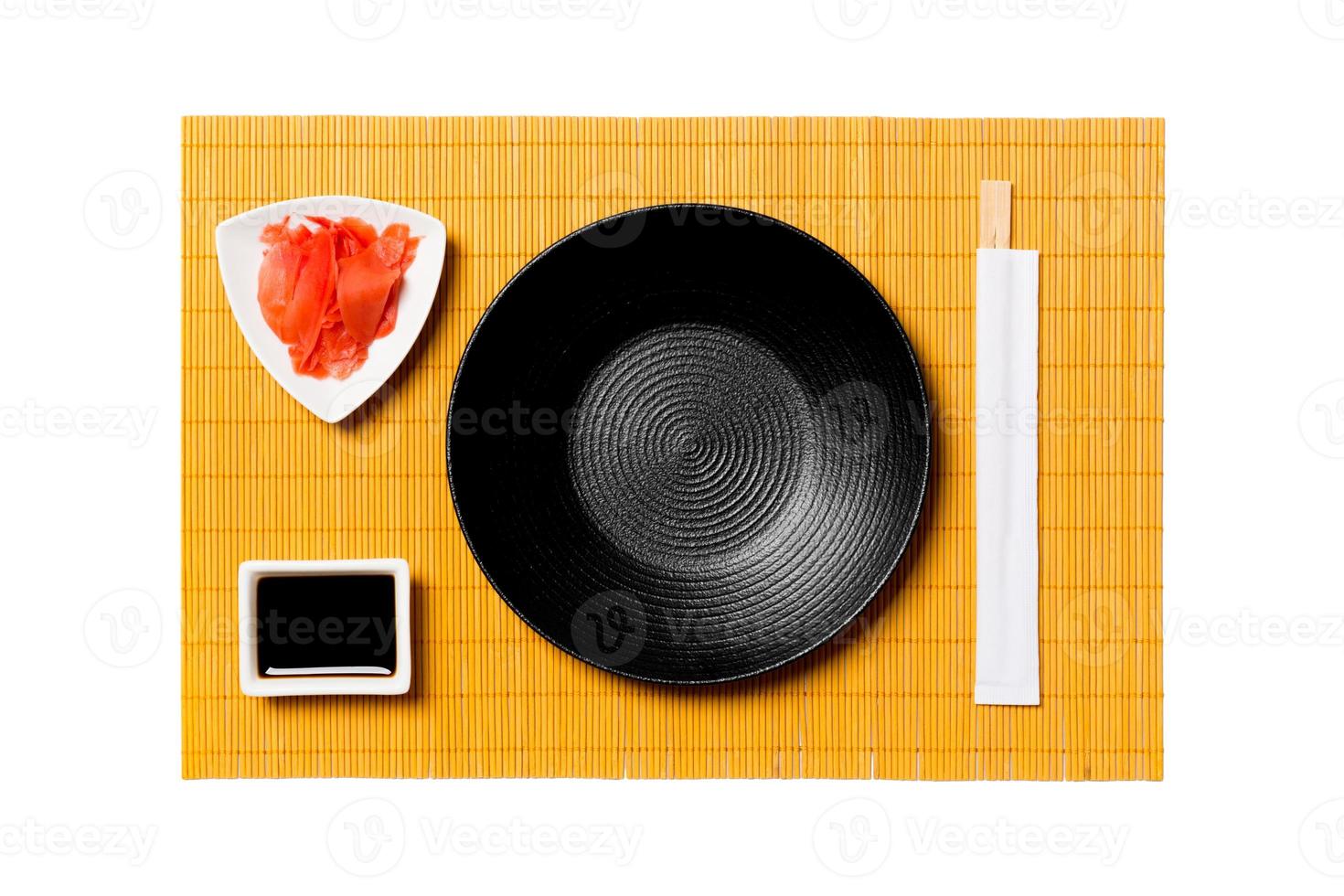 Empty round black plate with chopsticks for sushi and soy sauce, ginger on yellow bamboo mat background. Top view with copy space for you design photo