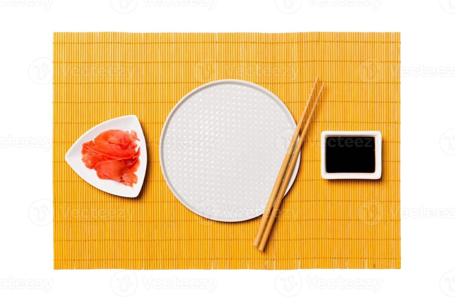 Empty round white plate with chopsticks for sushi and soy sauce, ginger on yellow bamboo mat background. Top view with copy space for you design photo