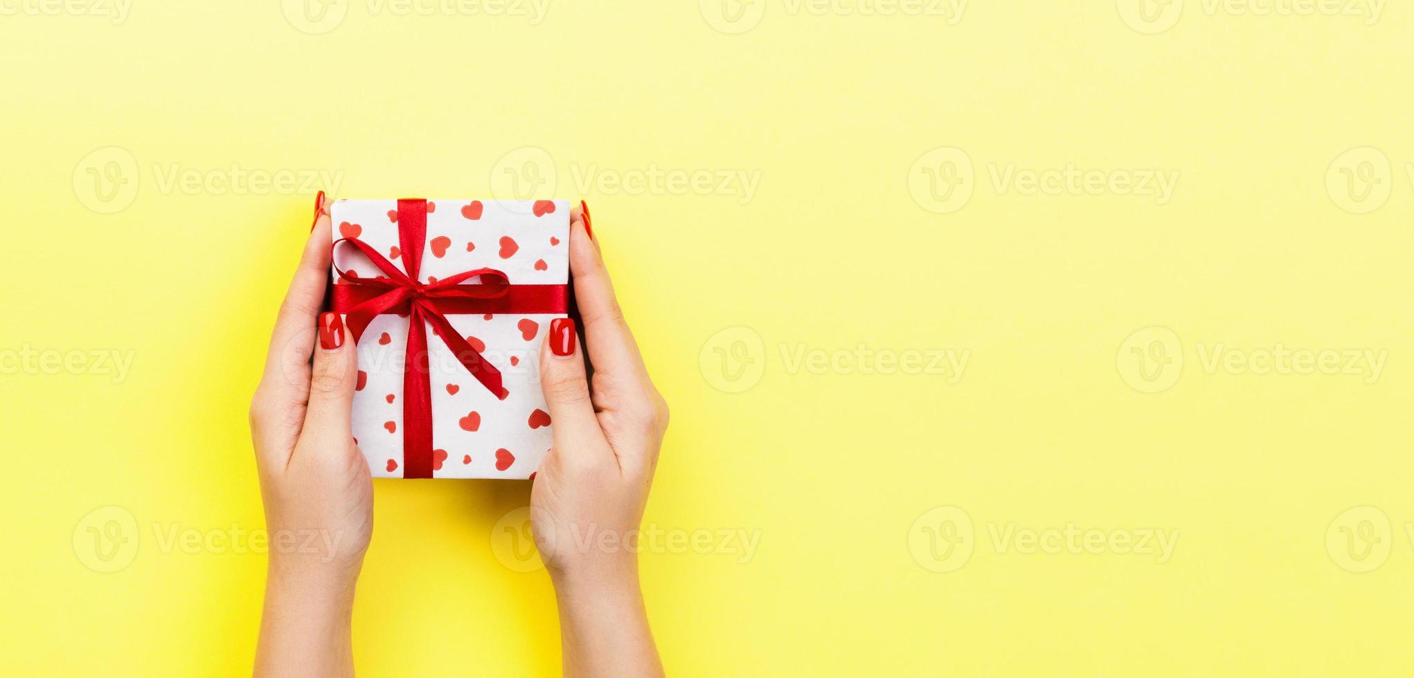 Woman hands give valentine or other holiday handmade present in paper with red ribbon. Present box, red heart decoration of gift on yellow table, Top view banner with copy space for you design photo