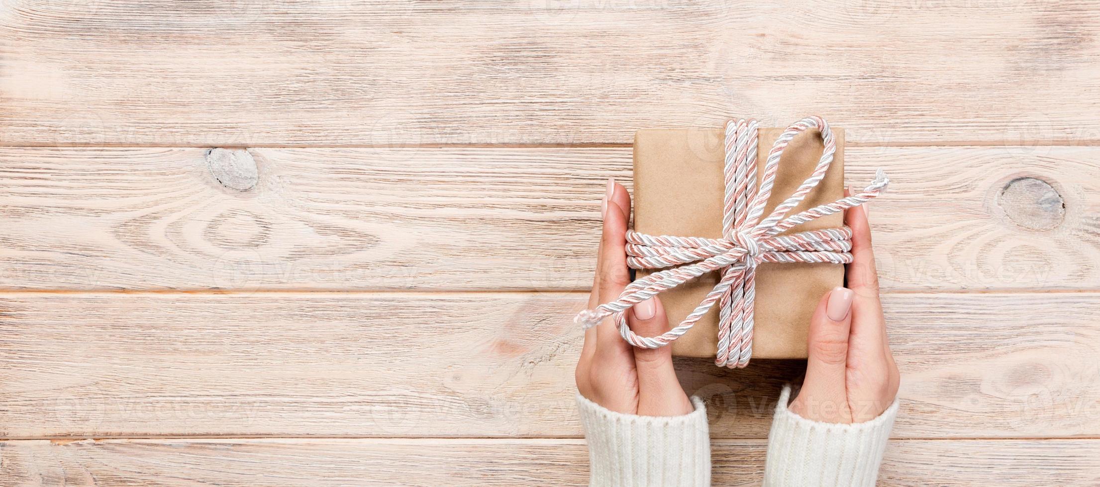 Woman hands give wrapped valentine or other holiday handmade present in paper with blue ribbon. Present box, decoration of gift on white wooden table, Top view banner with copy space for you design photo