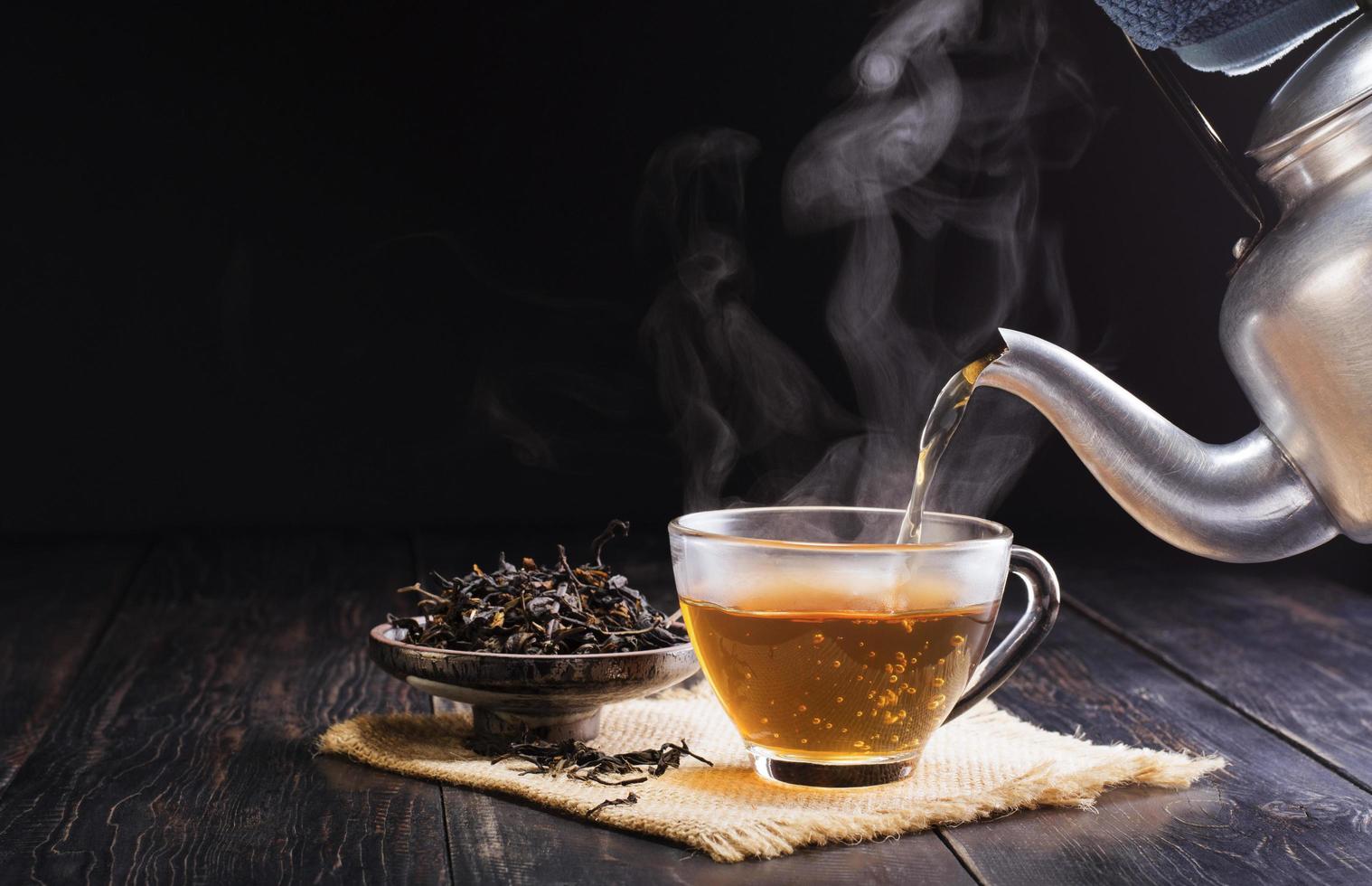 Pour the hot herbal teapot into a glass cup. Teacup is placed on a black wooden table on dark background. The sunlight of the new morning shines in the warm atmosphere. photo