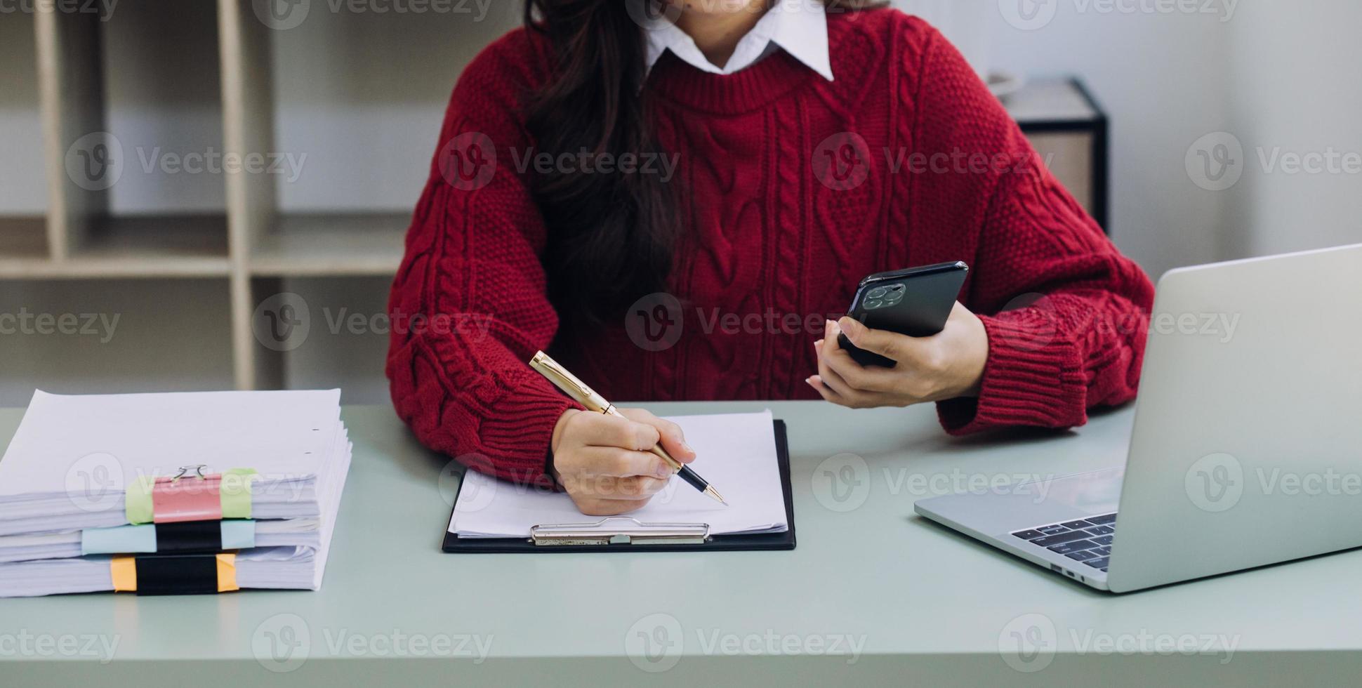 Young business woman sitting in office at table and using smartphone. On desk is laptop and tablet computer, on screen charts and graphs. Woman analyzing data. Student learning online. photo