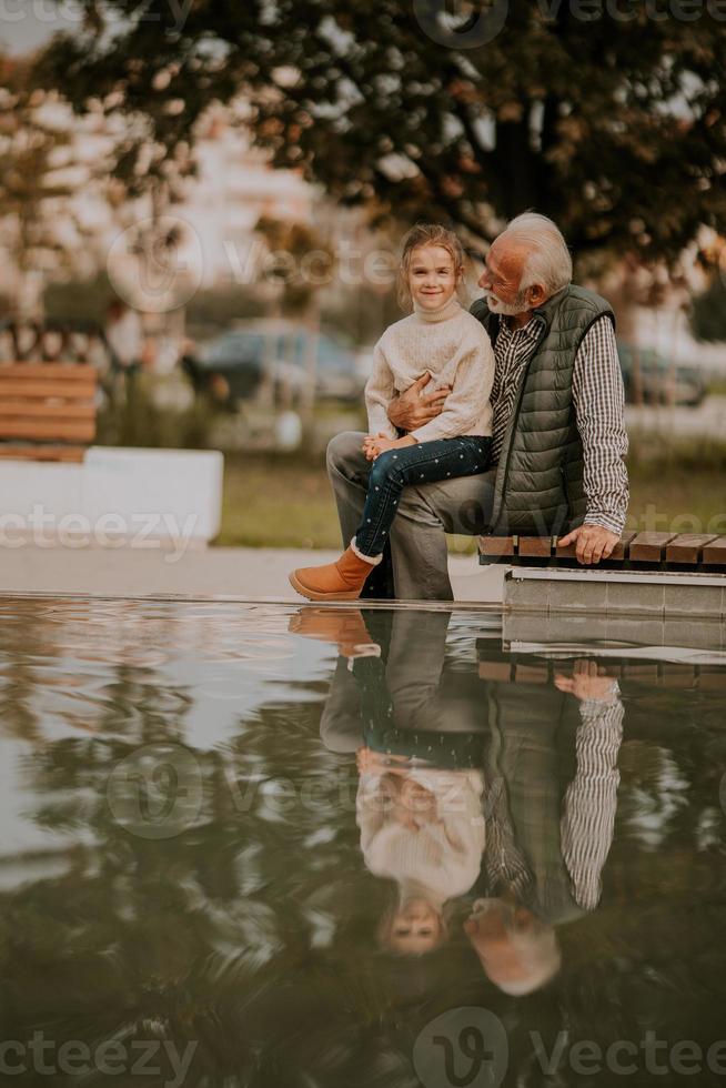 Grandfather spending time with his granddaughter by small water pool in park on autumn day photo