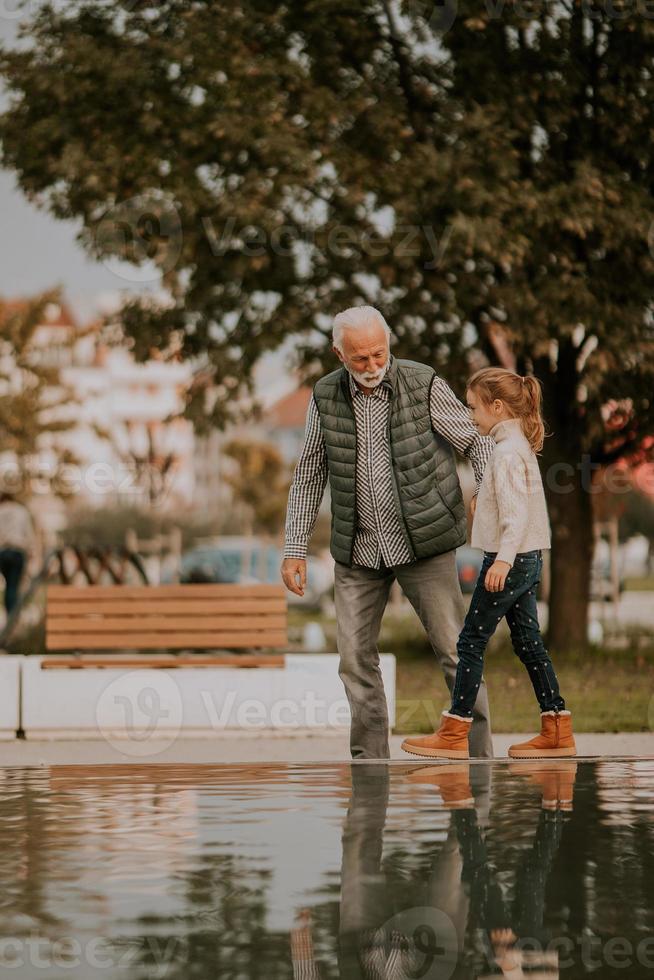 el abuelo pasa tiempo con su nieta junto a una pequeña piscina de agua en el parque el día de otoño foto