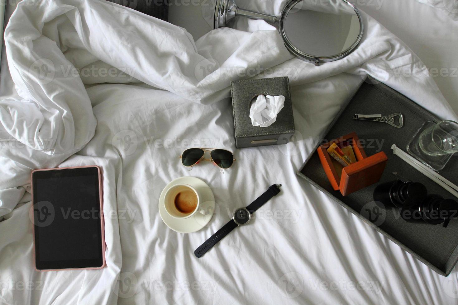 Top view of tablet, watch, black glasses, cup of coffee, gray box of napkin, bottle opener, wooden box and plastic conditioner shampoo and bath shower gel on white blanket. Working, relax and drink photo