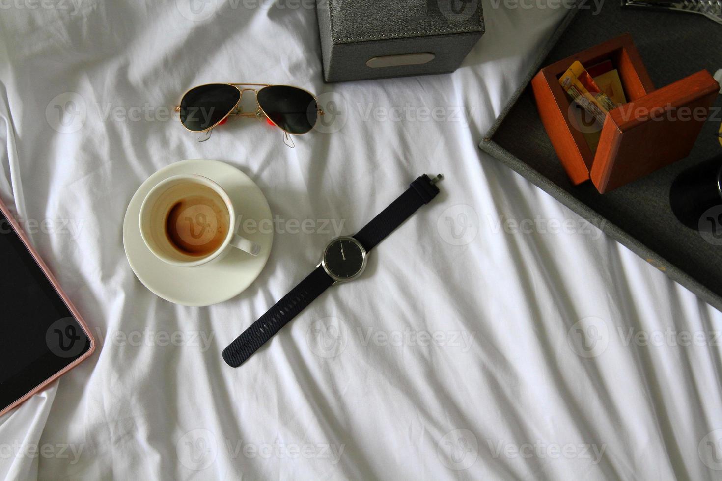 Top view of tablet, watch, black glasses, cup of coffee, gray box of napkin, bottle opener, wooden box and plastic conditioner shampoo and bath shower gel on white blanket. Working, relax and drink photo