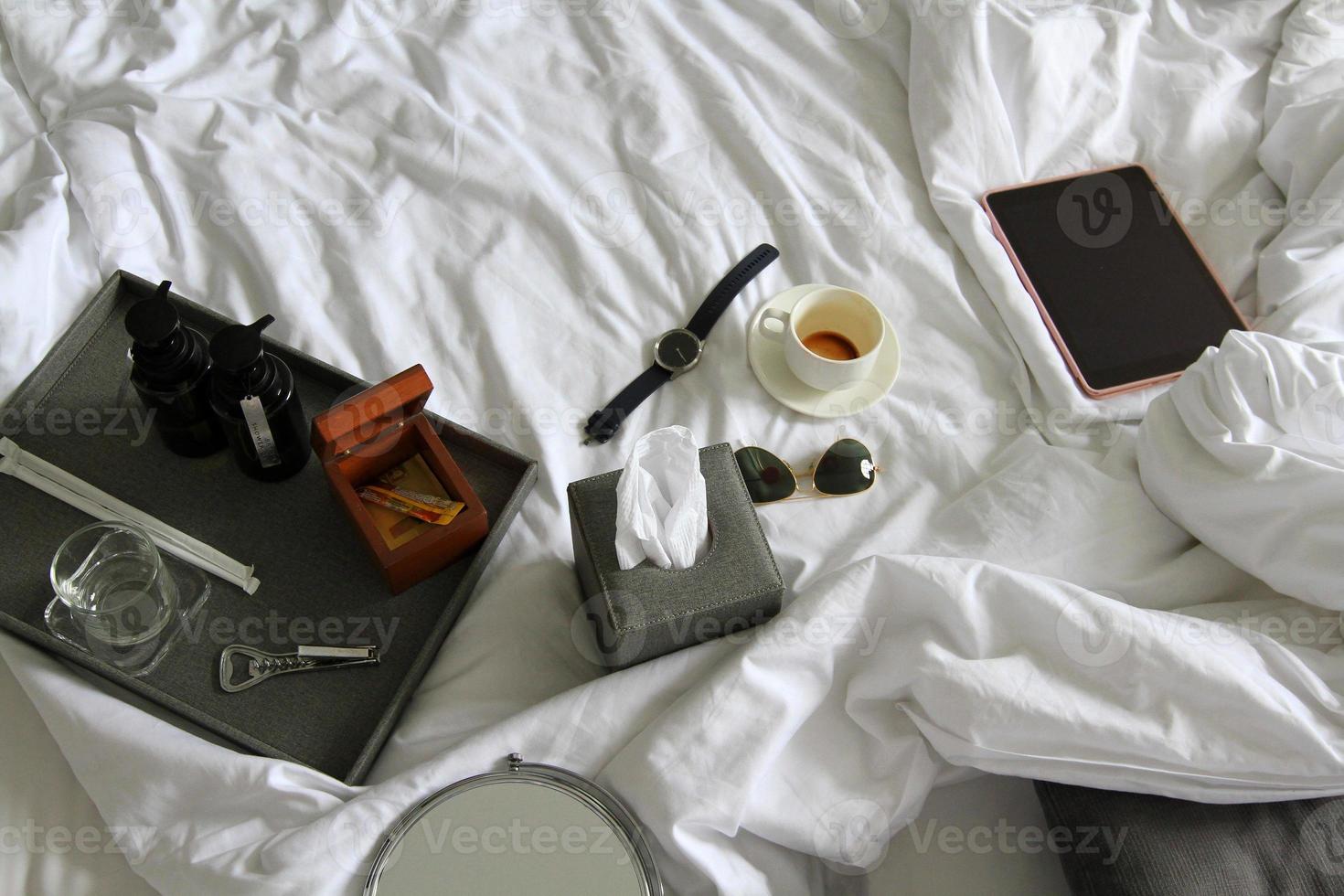 Top view of tablet, watch, black glasses, cup of coffee, gray box of napkin, bottle opener, wooden box and plastic conditioner shampoo and bath shower gel on white blanket with copy space. Object, photo