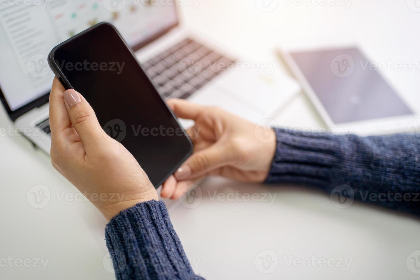 Woman is using a smartphone with a blank screen and a laptop on a nearby table. She is sitting in the office wearing a sweater. photo