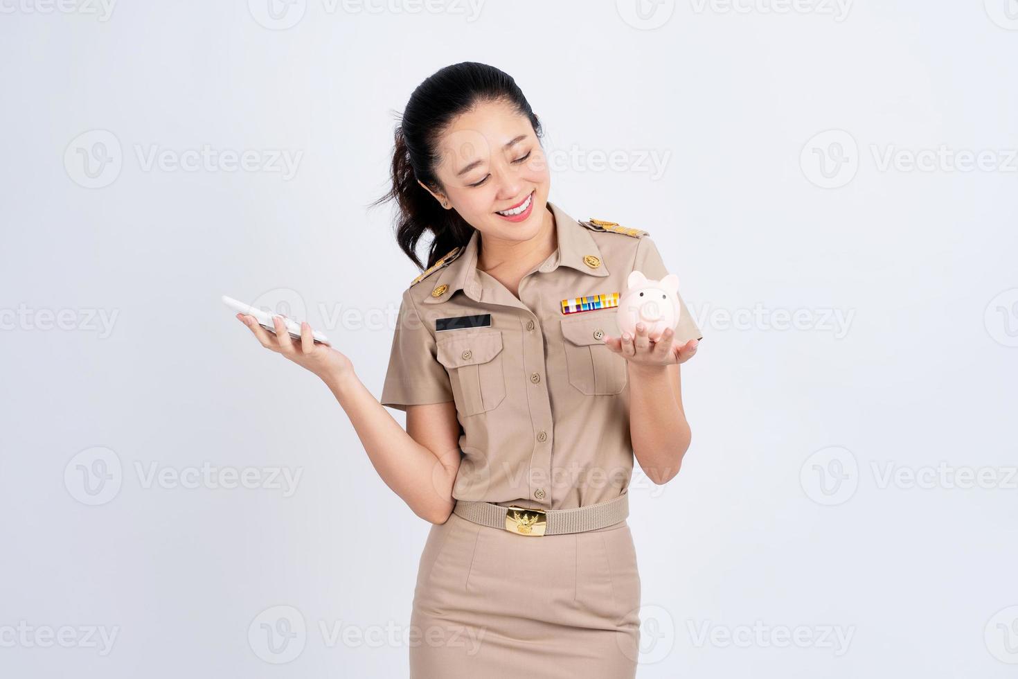 Portrait of excited Asian woman in brown uniform work clothes holding calculator and piggy bank isolated on white background, Business and financial concept photo