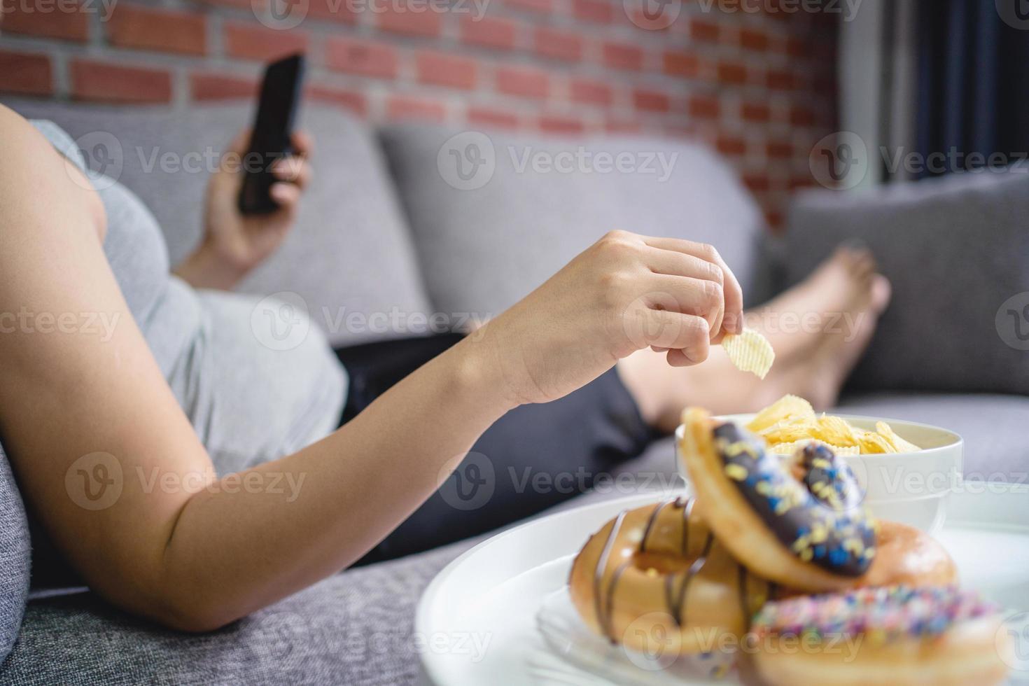 Fat woman lying on sofa holding smartphone and eating potato chips, overeating concept photo