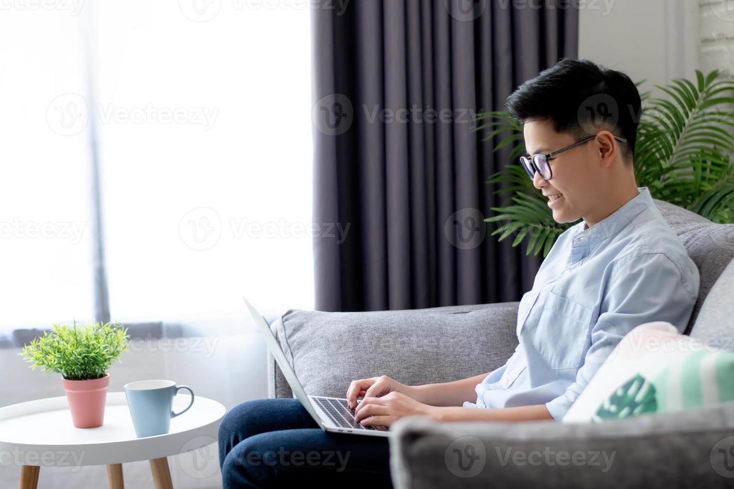 Side view of young Asian working near a large window and sitting on the sofa at home, With copy space. photo