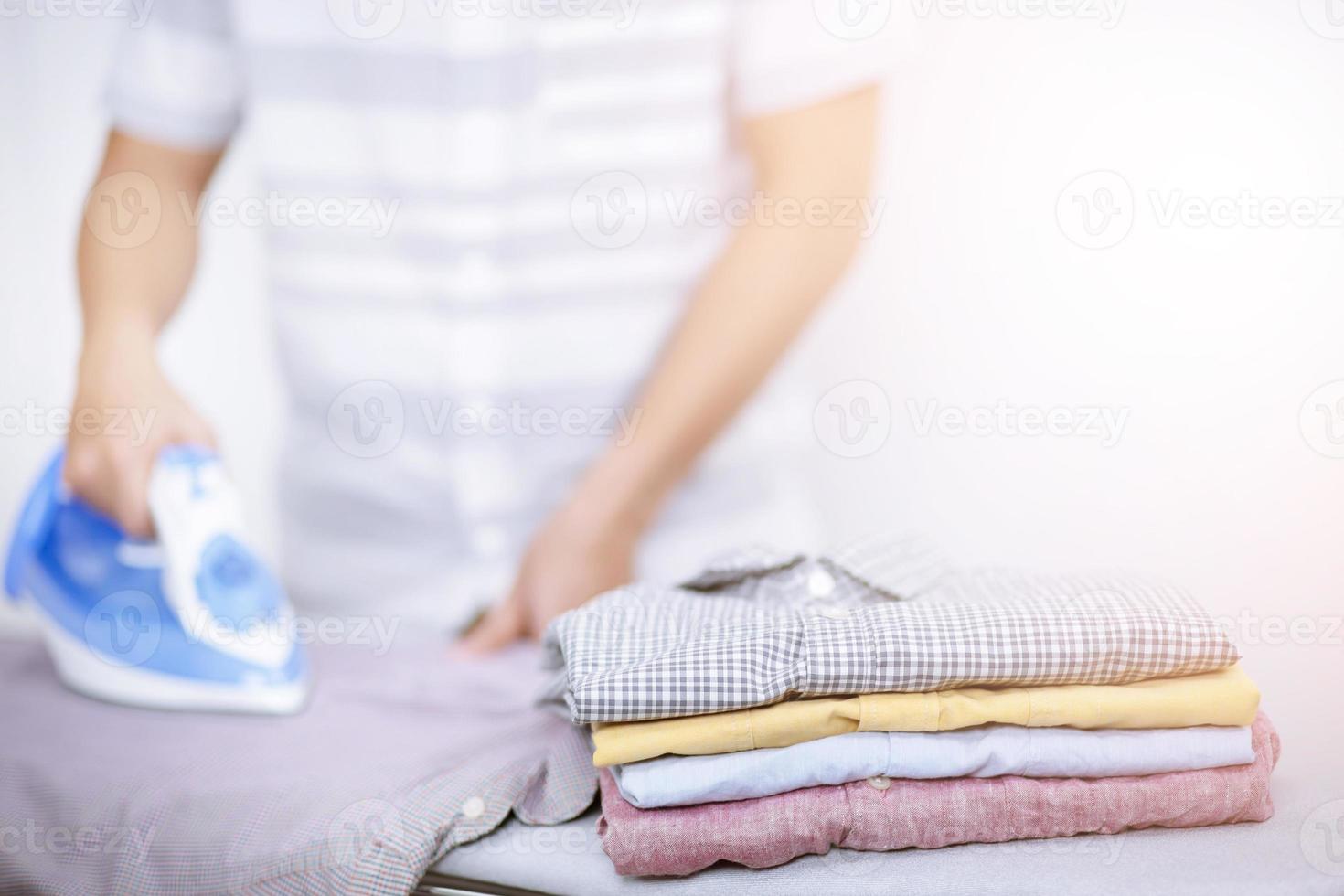 hombre en casa plancha electrica y pila de ropa variedad de colores. hombres planchando camisa en tabla de planchar con plancha azul humeante. tareas domésticas y concepto de hogar. foto
