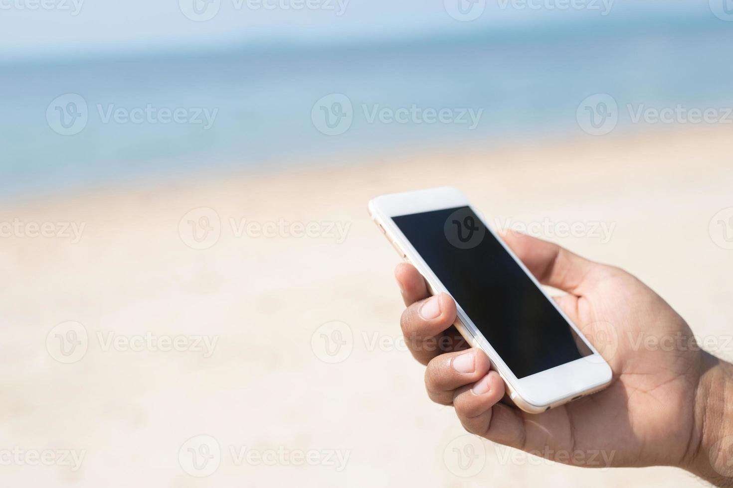 close up people man of hand holding white mobile smart phone of on the beach and sea background. with blank empty screen. empty space for text. Technology and travel holiday concept photo