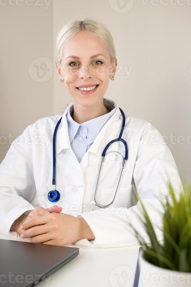 retrato vertical de una joven doctora caucásica sonriente sentada en un escritorio en un hospital o clínica privada con uniforme médico blanco en el lugar de trabajo, concepto de atención médica foto