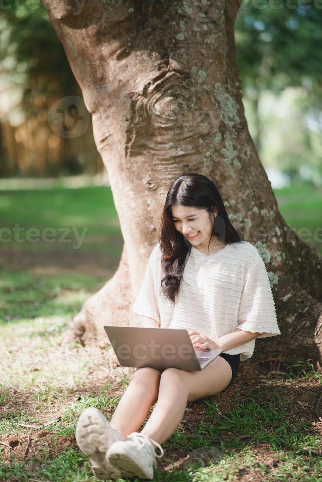 Asian woman sitting on picnic and lawn at park working on laptop. Asian female using laptop while sitting under a tree at park with bright sunlight. Work from anywhere concept photo