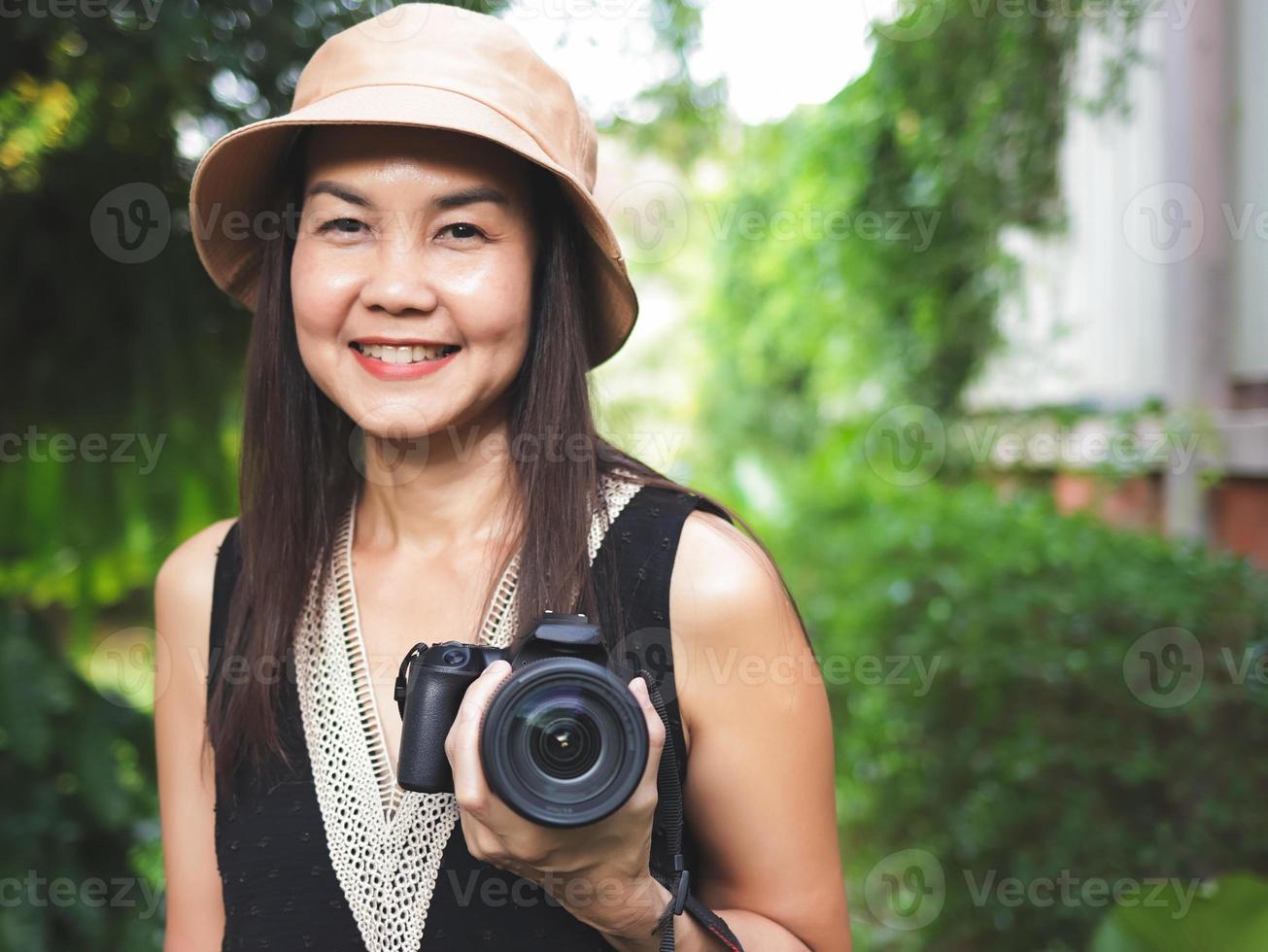 mujer asiática, con sombrero y blusa negra sin mangas, parada en el jardín y sosteniendo una cámara dslr, sonriendo alegremente. foto