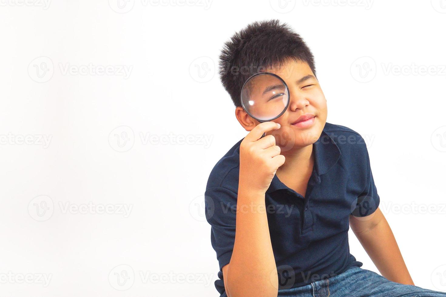 Boy holding a magnifying glass looking to camera with fun on white background. Curiosity, research and explore concept. photo