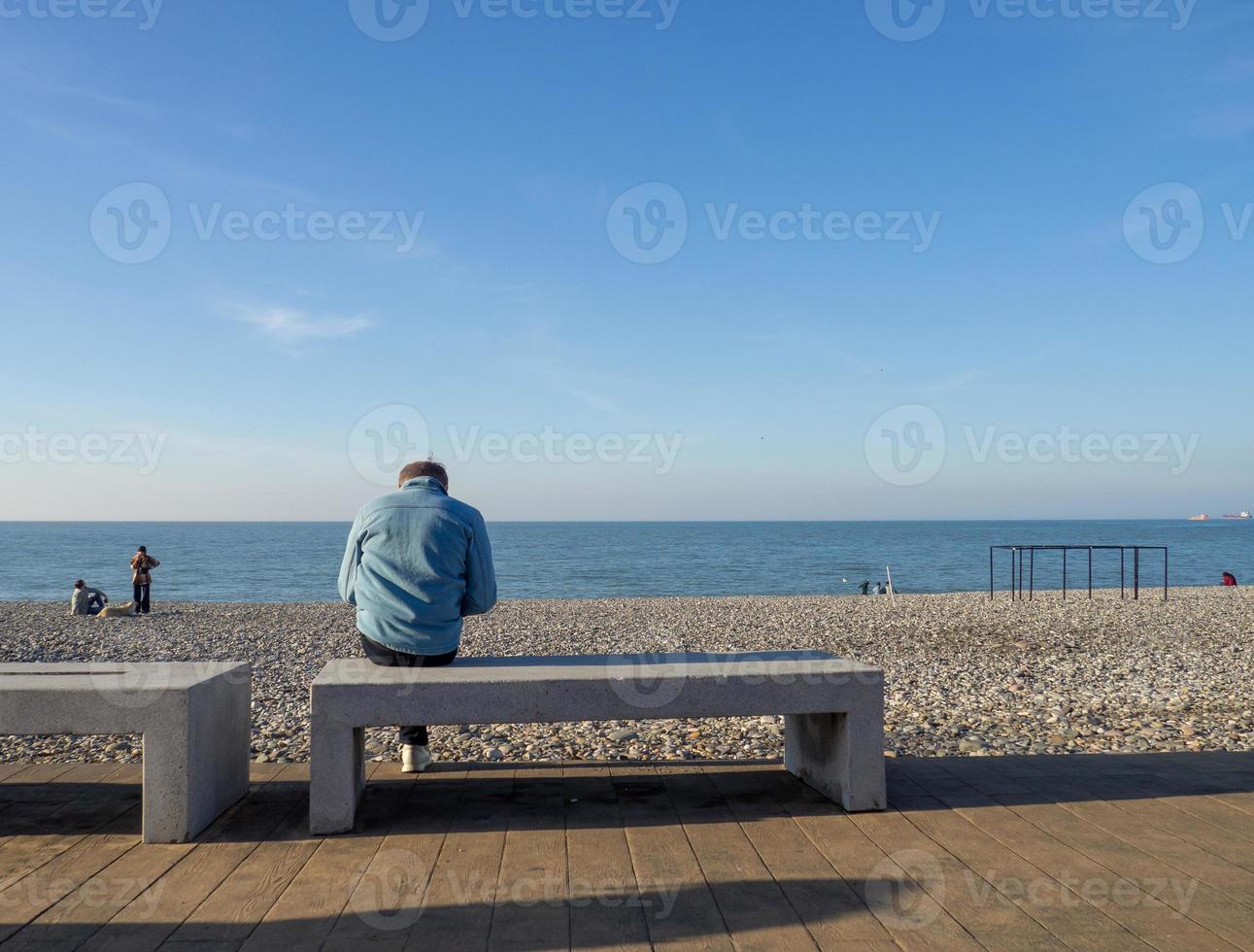 Man on a bench by the sea. A man with a phone is sitting by a pebbly beach. Vacation on the beach. Relaxation at sea. Rocky seashore. Pebble beach. Boats on the water. photo