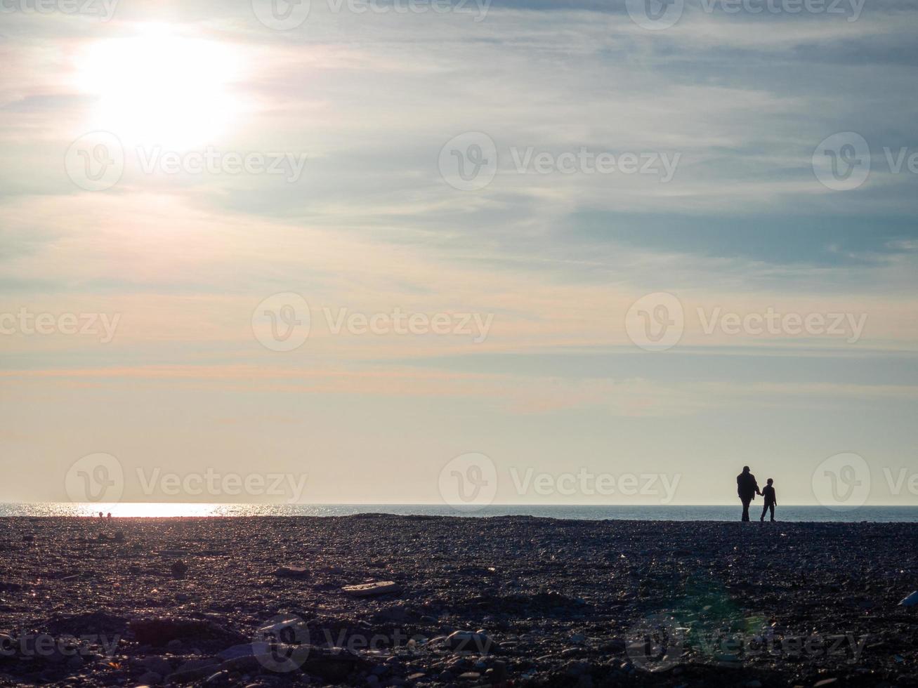 Father and son on a spring pebble beach at sunset. Silhouettes of people on the shore. Vacation on the beach. Rest on the sea. Rocky shore. photo