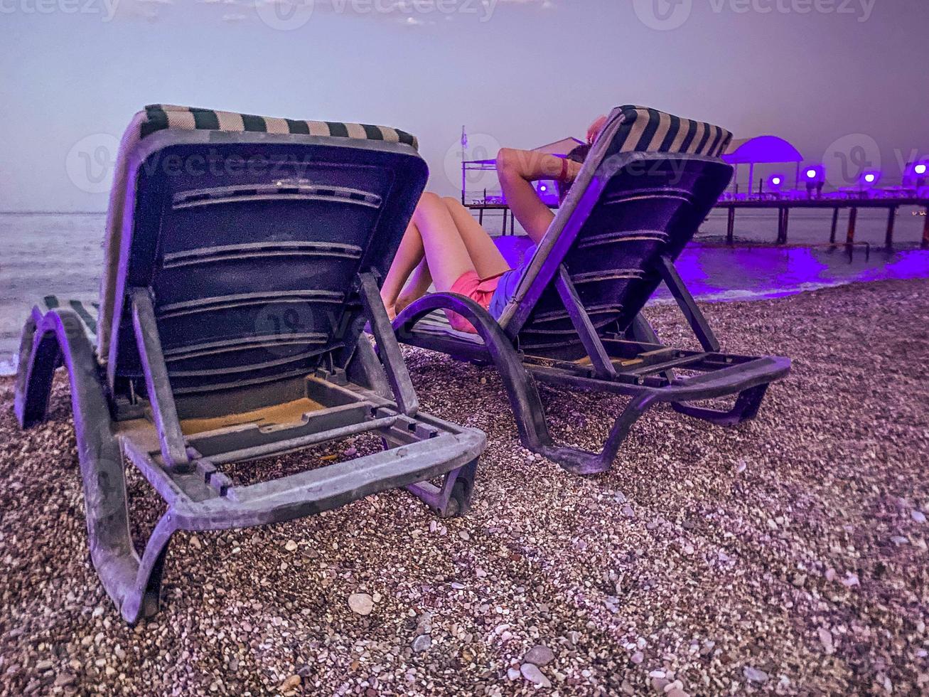 hot, tropical country with sea and mountains. on the beach there are pebbles and small stones. there is a green sunbed on the ground, a girl in bright shorts is resting on it photo