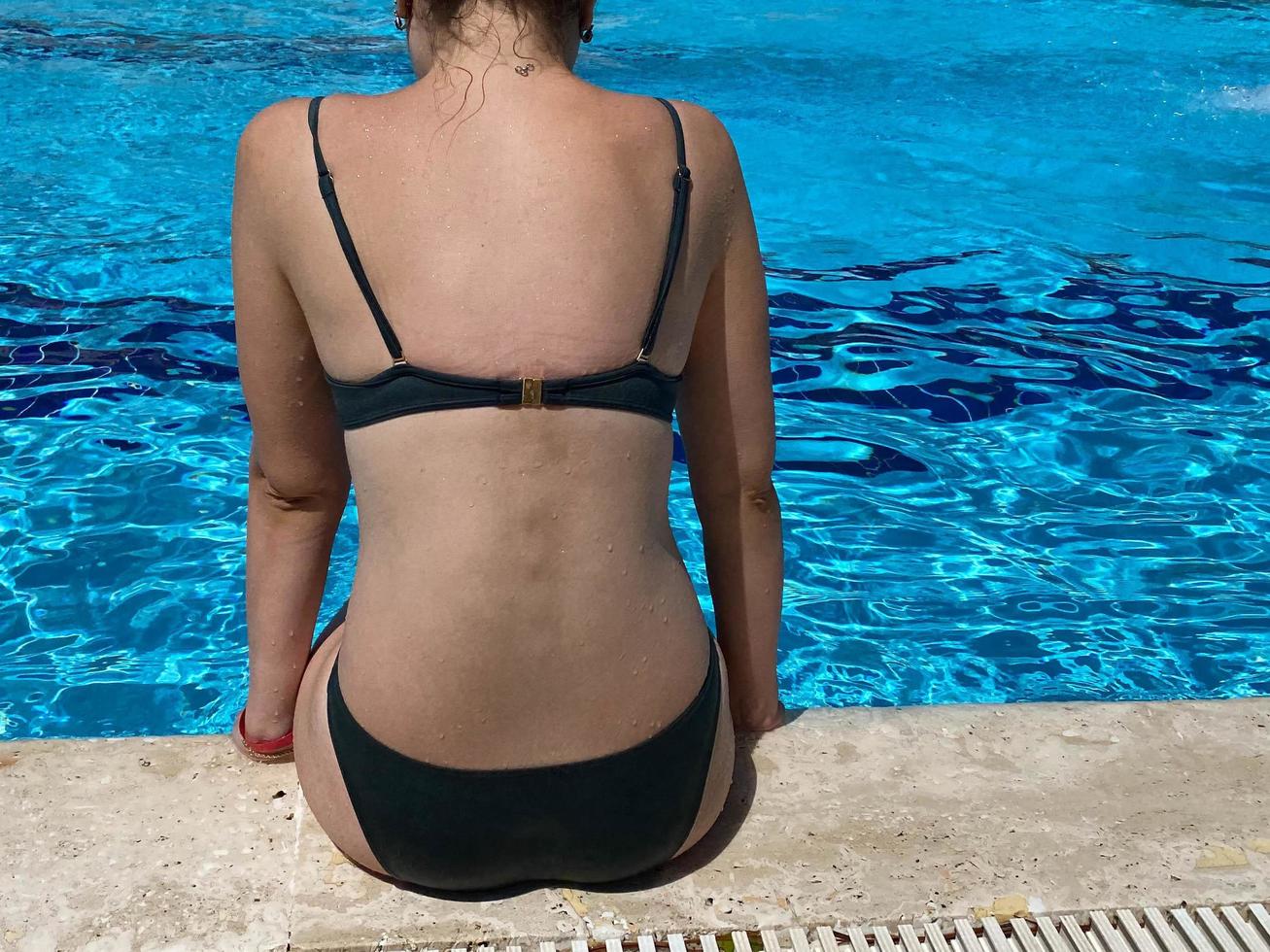 Sexy fit european woman with two braids in tiny black bikini sits on edge of swimming pool back to camera, tropical background, sunset light photo