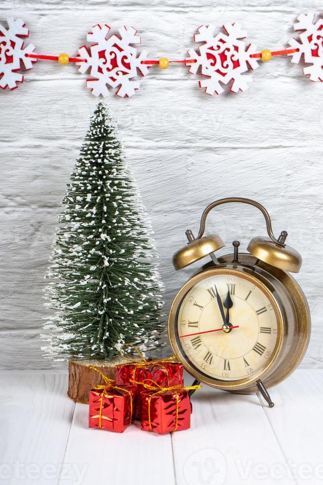 Decorative christmas tree, gift boxes and alarm clock on white wooden background. photo