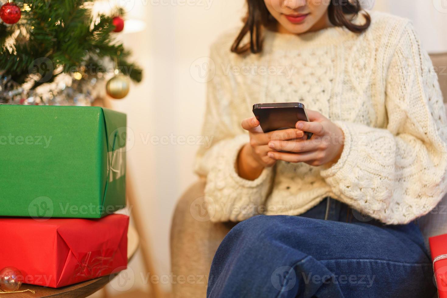 concepto de celebración de navidad, joven asiática sentada en la sala de estar y charlando en el teléfono inteligente foto