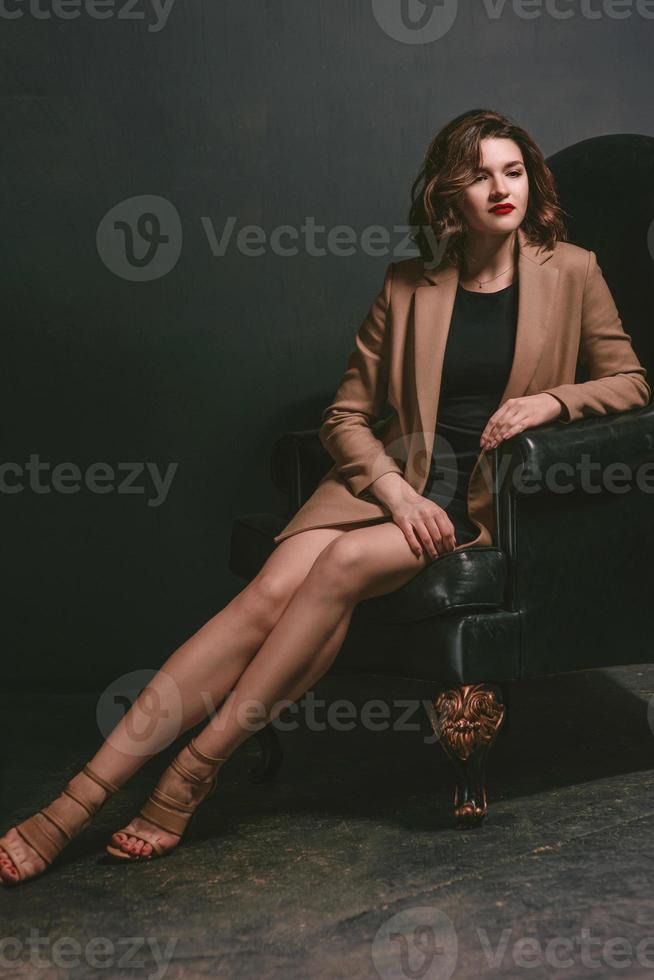 portrait of young, beautiful sad woman with short brown hair with stylish make up in black dress and beige jacket in armchair on dark background photo