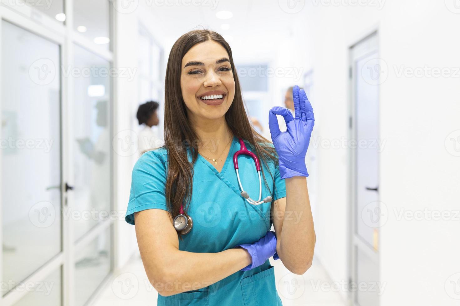 Young female medical doctor working at the hospital and medical staff, she is showing OK sign. Nurse stands in a walkway and smiles with her arms folded. She is wearing scrubs and a stethoscope. photo