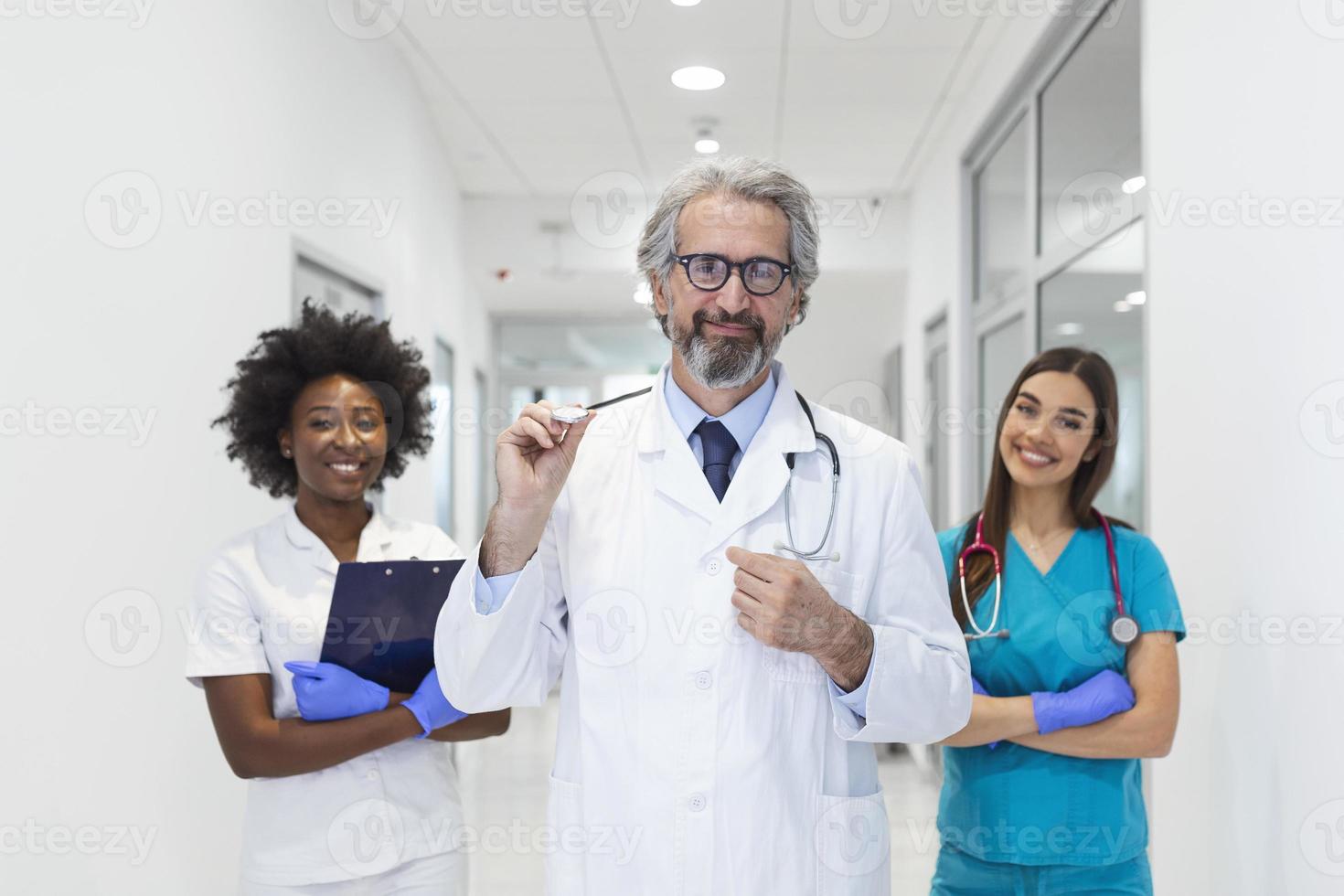 A multi-ethnic group of medical staff . They are dressed in medical scrubs and white lab coats with stethoscopes around their necks. Some standing in the back. The focus is on an mature man in front photo