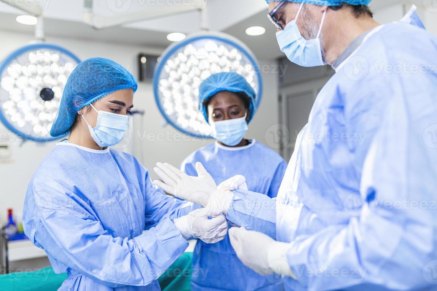 joven enfermera con máscara protectora y ropa de trabajo que ayuda al cirujano con guantes mientras ambos se preparan para la operación foto