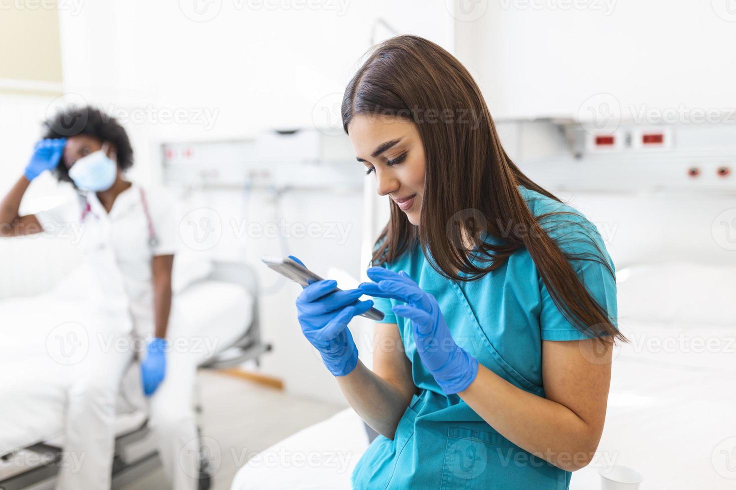 An exhausted female nurse and a african american doctor sits on a bed in a hospital during a shift break. She daydreams while holding his smartphone. photo