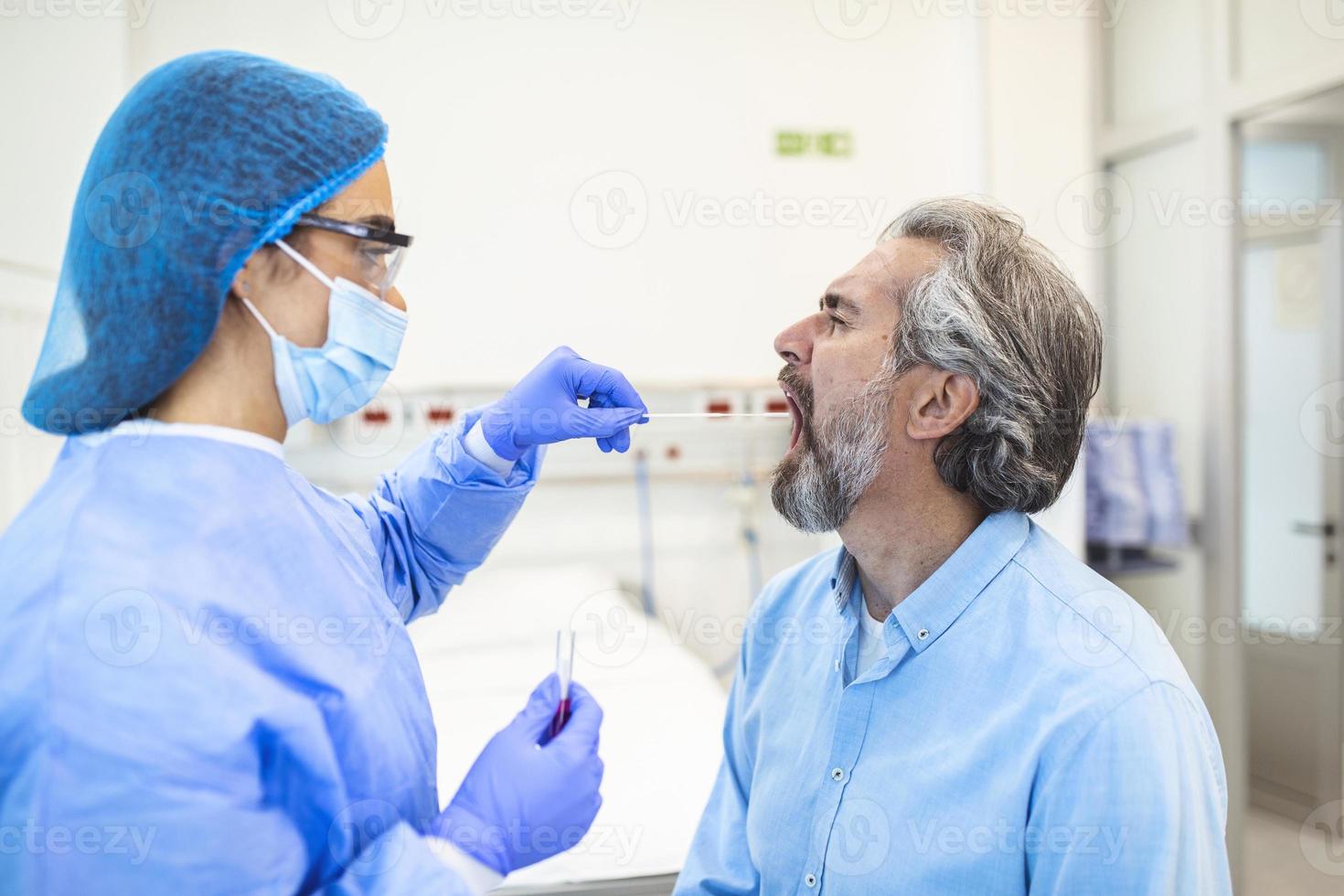 un hombre caucásico maduro en un entorno clínico siendo examinado por un trabajador de la salud con atuendo protector para determinar si ha contraído el coronavirus o covid-19. prueba pcr foto
