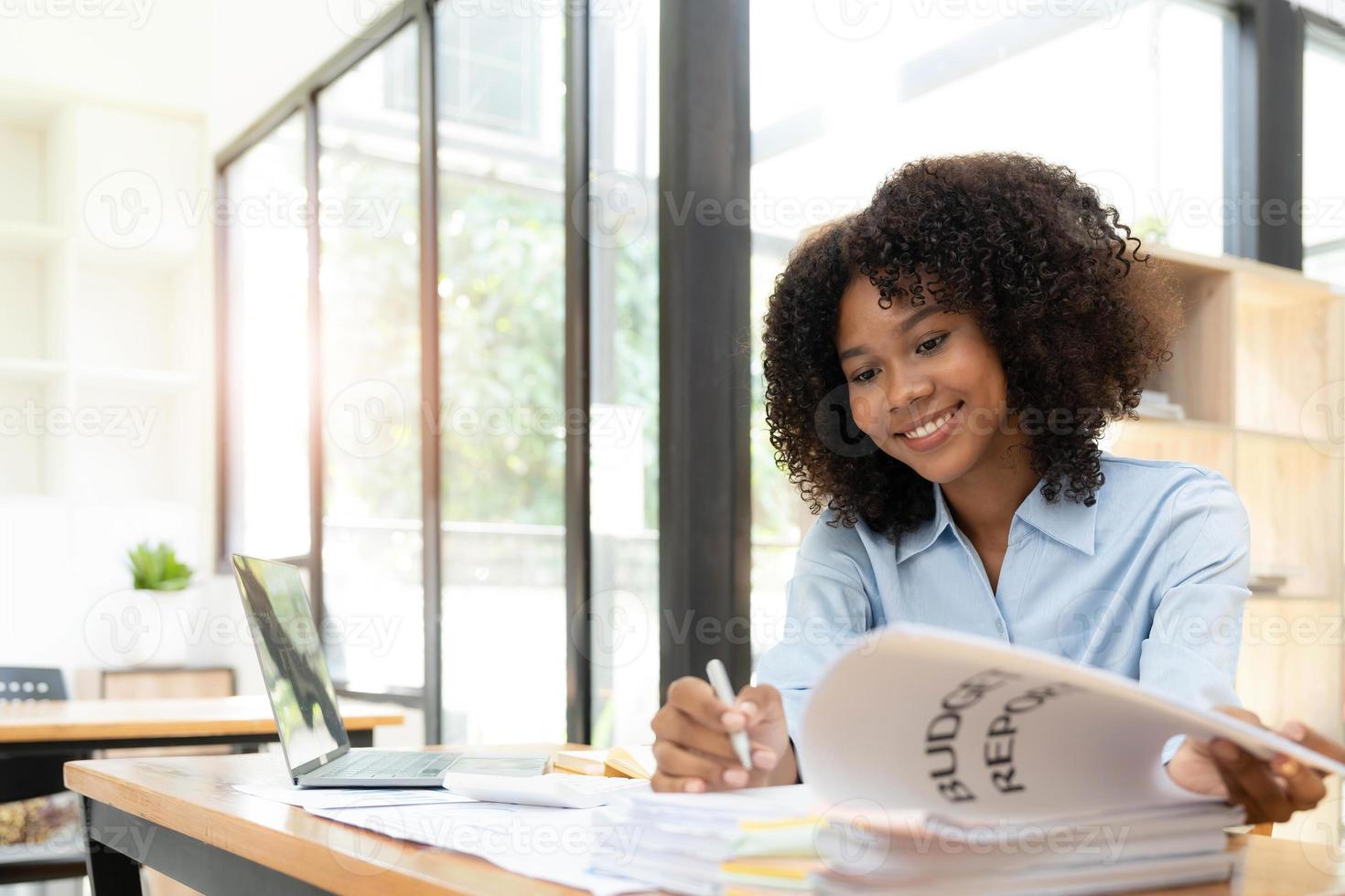 sonriente joven empresaria africana sentada en un escritorio en su oficina trabajando en línea con una laptop foto