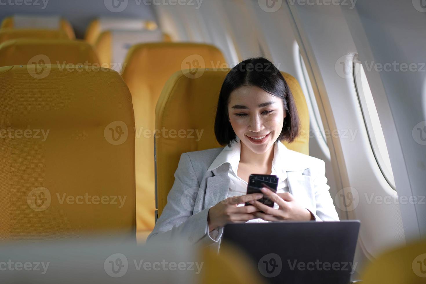 Asian woman passenger sitting in airplane near window and reading news from social networks or using travel applications in smartphone photo