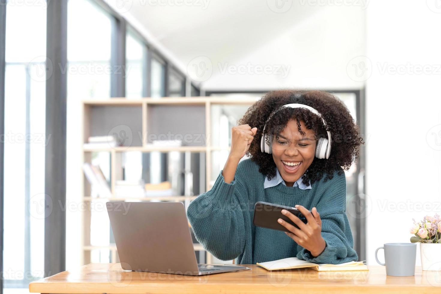 Photo of cheerful joyful mixed-race woman  playing games and wearing wireless headphones at home  Play game on your smartphone that wins the moment of victory. Fun and enjoyable vacation