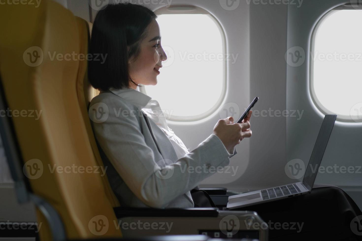 Asian woman passenger sitting in airplane near window and reading news from social networks or using travel applications in smartphone photo