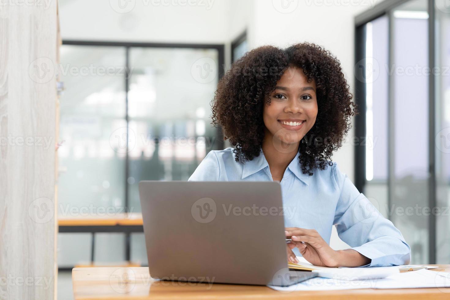 Smiling young African female entrepreneur sitting at a desk in her home office working online with a laptop photo