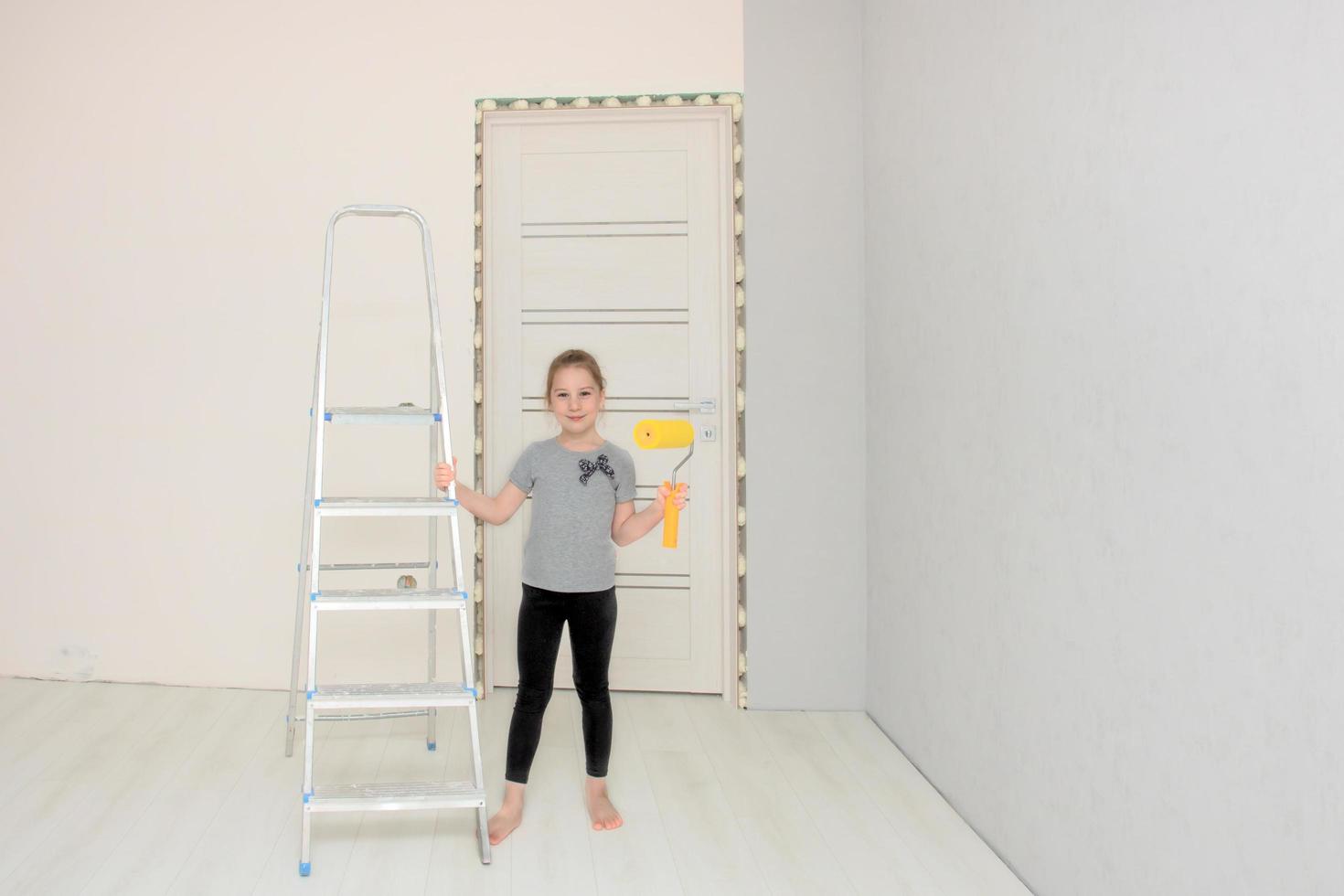 cute girl in the room where the renovation is underway stands next to the stepladder and holds a paint roller in her hand photo