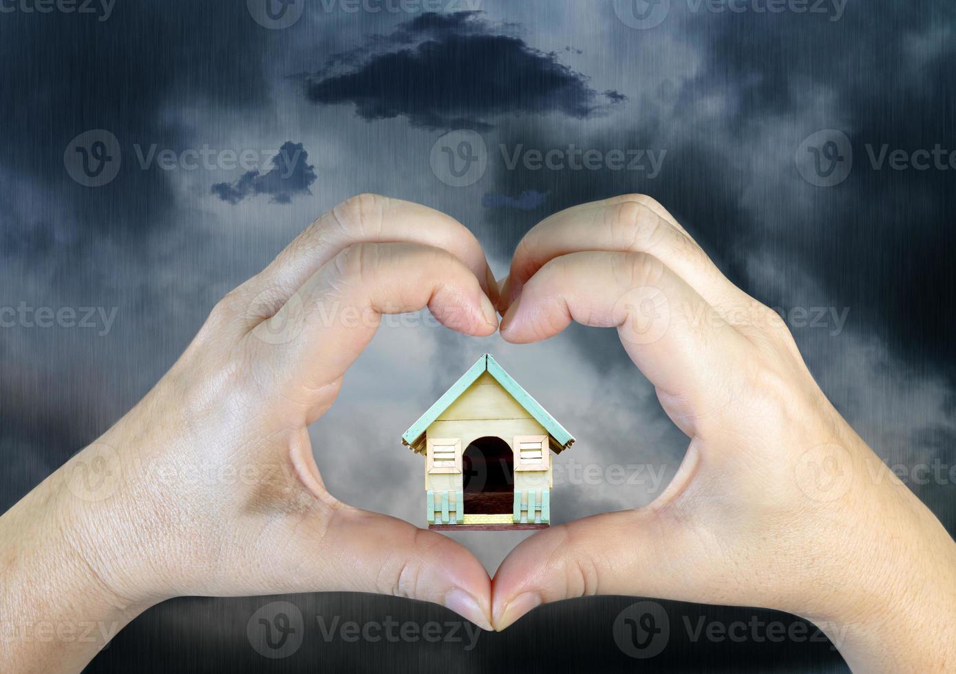 person hands making a heart shape with wooden house on rain clouds background photo