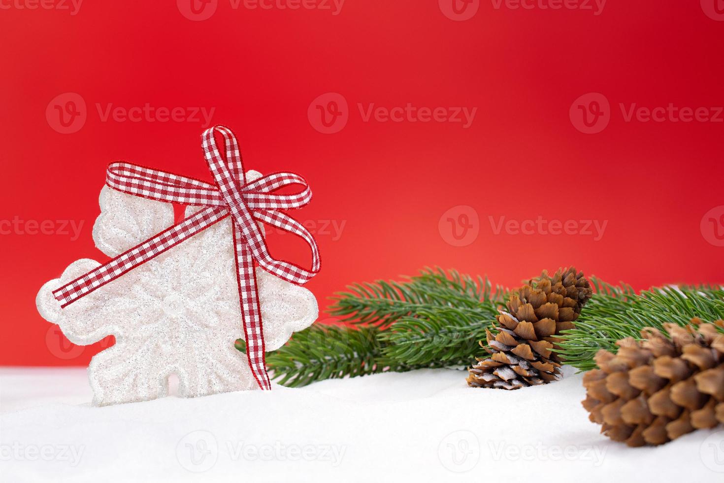 White shiny snowflake with bow, fir branches and cones on snow, red background behind. Christmas, New Year, winter. Copy space photo