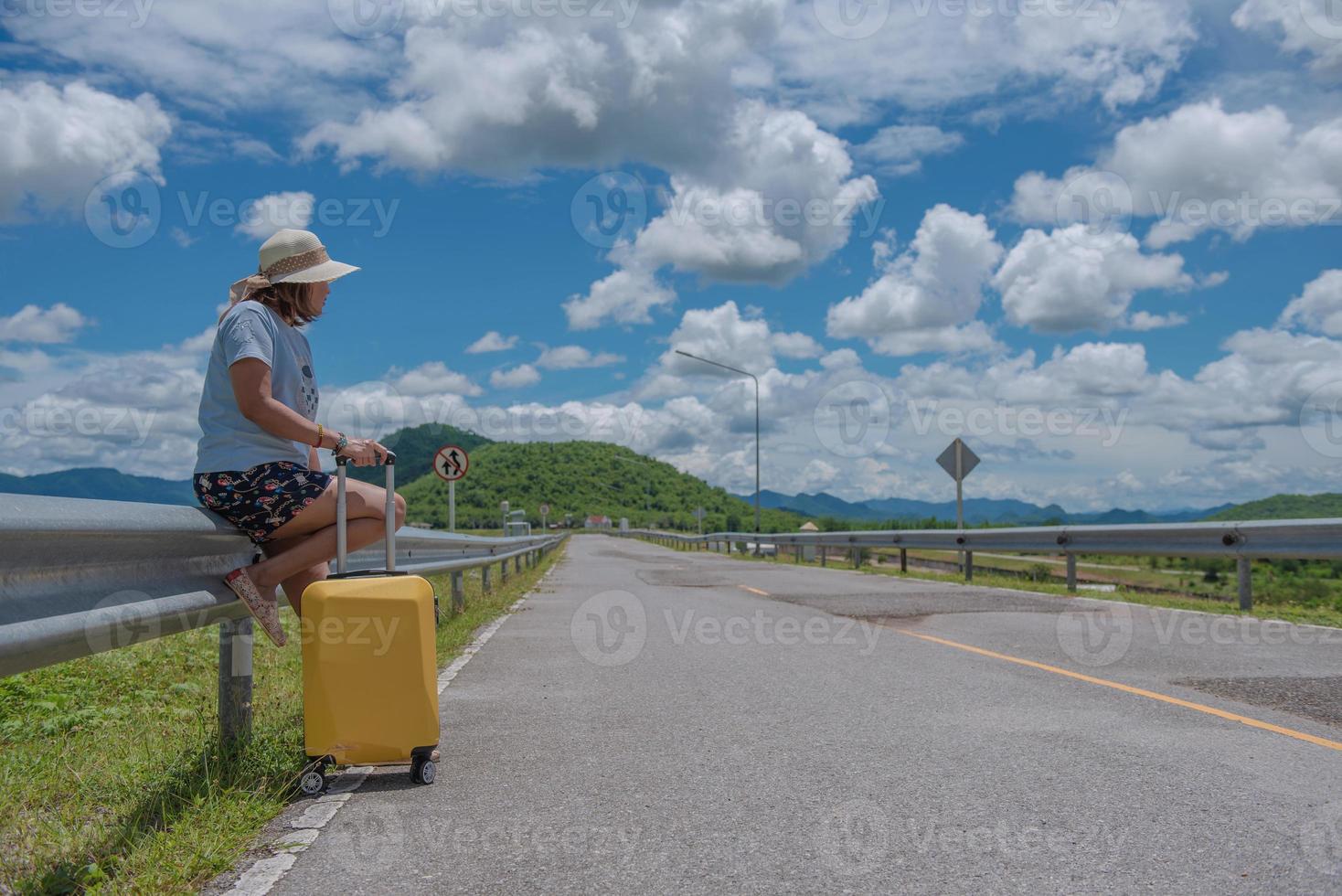 Hitchhike Asian woman on a road with traveling suitcase waiting for a car on beautiful natural landscape with blue sky, sunny day travel concept. photo