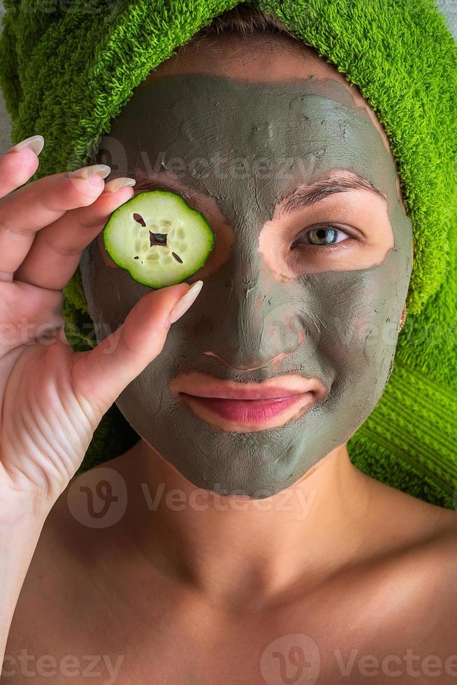 Beautiful young woman with facial mask on her face holding slices of cucumber. photo