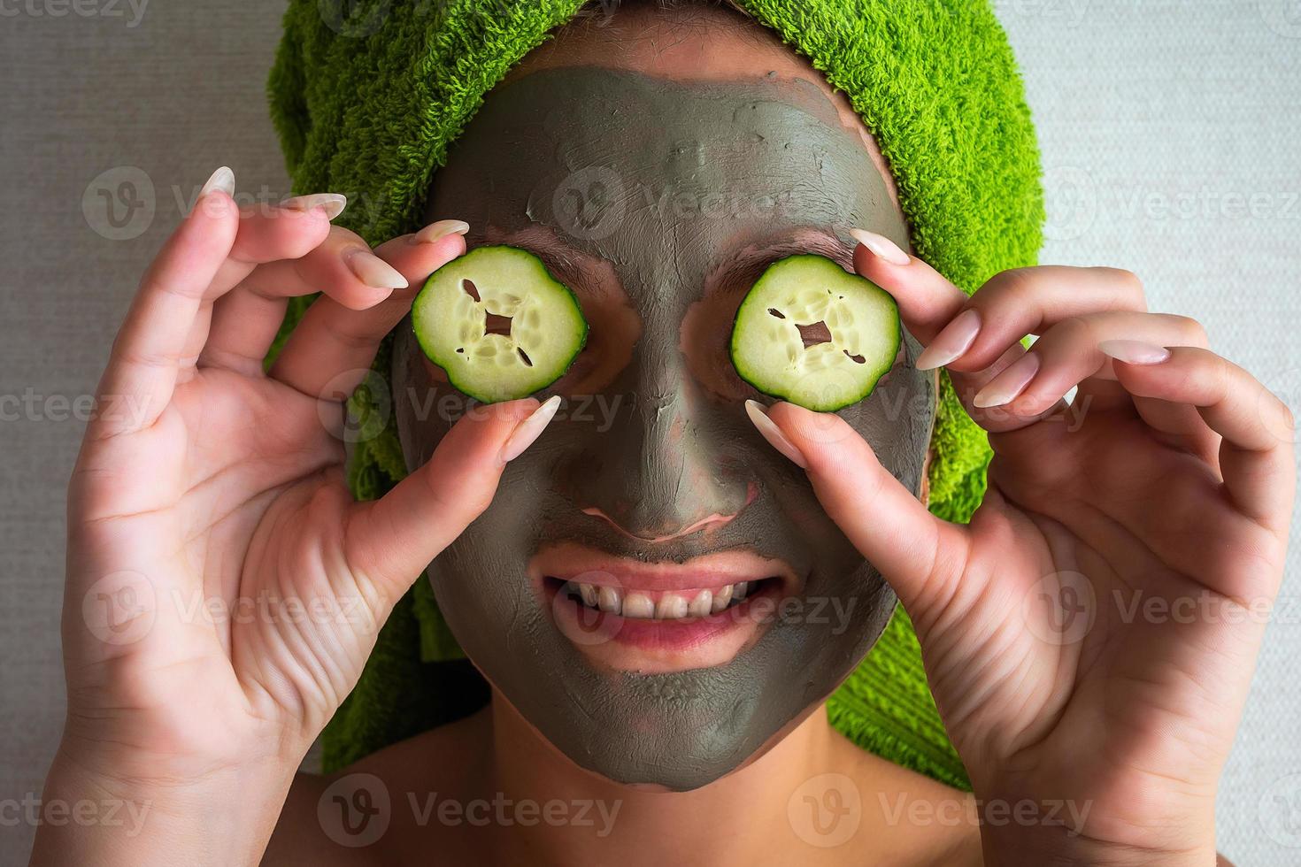 Beautiful young woman with facial mask on her face holding slices of cucumber. photo