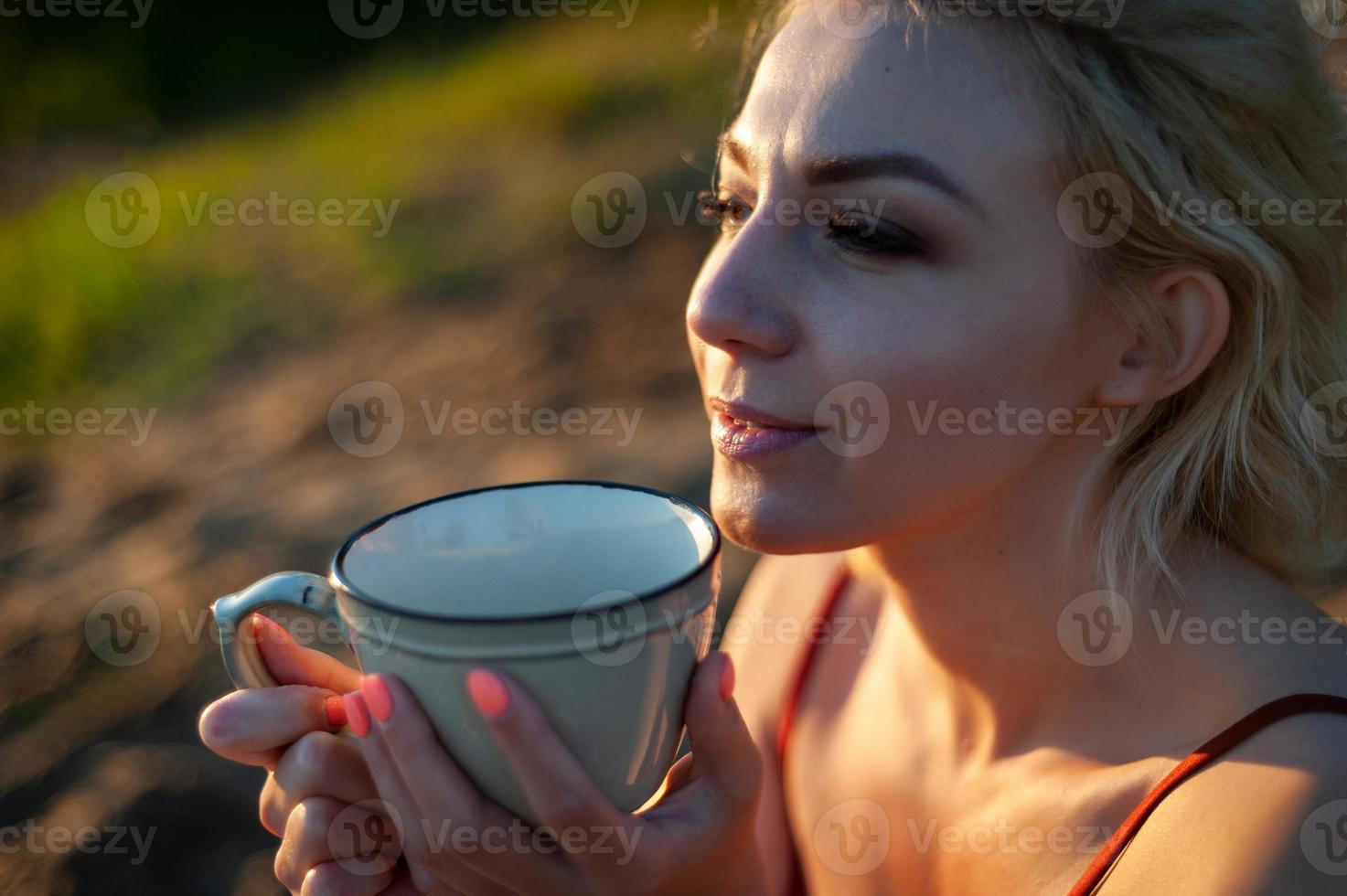 A beautiful girl, covered with a plaid in the evening at sunset, holds a mug of tea with two hands against the sea. photo