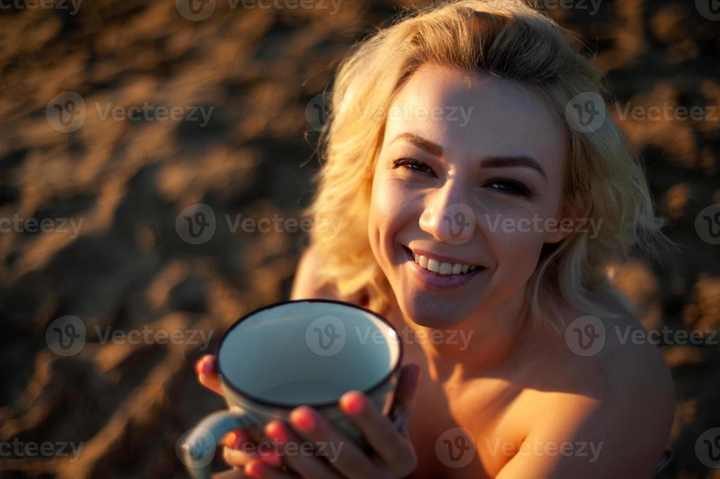 A beautiful girl, covered with a plaid in the evening at sunset, holds a mug of tea with two hands against the sea. photo